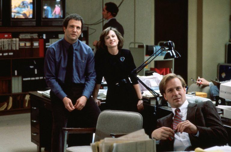 Still from the film Broadcast News. Albert Brooks and Holly Hunter lean against a desk, staring directly ahead as if watching television. William Hurt is seated in front of them, also staring, transfixed.